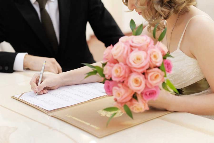 כלה מחזיקה זר פרחים חותמת על הסכם והחתן לצידה